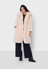 Gewatteerde mouwloze jas met een luxe, lichtroze nepbont kraag. Voorzien van zijzakken en een crèmekleurige buitenlaag, gecombineerd met zwarte wijde broek.