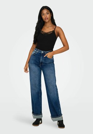 Jonge vrouw staand met hand in de zak, draagend een zwart tanktop met kanten rand, hooggetailleerde blauwe wijdere broek met omslagen en zwarte sneakers.