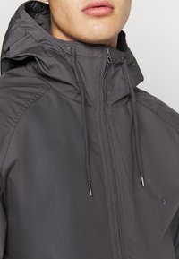 Primer plano de una persona que lleva una chaqueta de cremallera gris oscuro con capucha y cordones, mostrando solo los labios y la parte inferior de la cara.