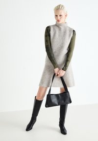 Abito grigio lavorato a maglia con colletto alto e orlo svasato, abbinato a un maglione verde a maniche lunghe e una borsa in pelle nera. Stivali neri alti fino al ginocchio.