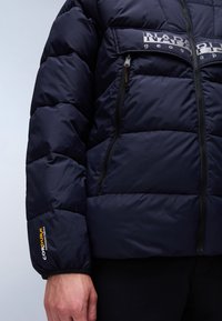 Námořnická puffer bunda s prošívanou texturou, vybavená zapínacími kapsami a logem ‚NAPAPIJRI‘ na hrudi. Značka Cordura na manžetě.