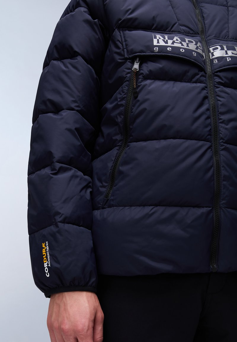 Marineblauwe pufferjas met gewatteerde textuur, voorzien van ritszakken en een 'NAPAPIJRI' logo op de borst. Cordura-branding op de manchetten.