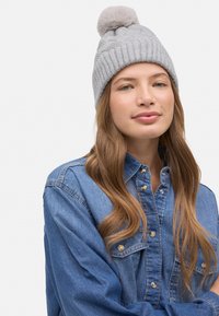 Gorro de punto gris con textura acanalada y un suave pompón en la parte superior, combinado con una camisa vaquera azul con botones de presión.