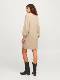 Beige kötött ruha laza fazonnal, hosszú puffos ujjakkal és oldalsó hasítékokkal, fekete cowboy csizmával párosítva. Simább textúrájú.