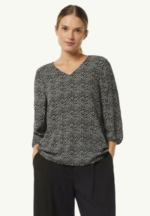 Black and white chevron-patterned blouse with a V-neck, three-quarter sleeves, and a relaxed fit, worn over black trousers.