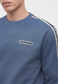 Мъж, носещ син суитшърт с лого EA7 Emporio Armani на гърдите и лента с марка по рамото и ръкава.