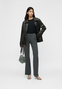 Γυναίκα με μαύρο δερμάτινο μπουφάν, μαύρο τοπ, γκρι παντελόνι, κρατώντας τσάντα με σχέδια και φορώντας πέδιλα με ανοιχτά δάχτυλα και τακούνι, στέκεται σε λευκό φόντο.