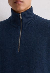 Dunkelblaues Sweatshirt mit einem gerippten hohen Kragen, ausgestattet mit einem silbernen Reißverschluss und einem strukturierten Stoff mit dezenten Sprenkeln, der ein gemütliches Erscheinungsbild erzeugt.