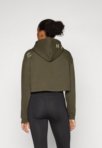 Hoodie verde-oliva cortado com capô e bolso frontal. Apresenta texto impresso no ombro direito. Combinado com leggings pretas.