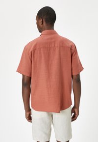 Camisa de manga corta en color óxido, de tejido liviano; tiene cuello, dobladillo recto y corte holgado. Se lleva con pantalones cortos beige.