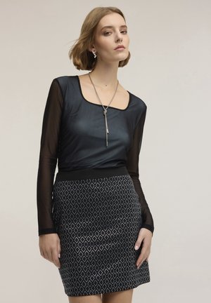 Ung kvinna som bär en svart genomskinlig långärmad topp och en svart kjol med silverfärgade diamantmönster, poserande mot en enfärgad bakgrund.