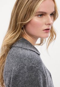 Γκρι μάλλινο πουλόβερ με υφή, με γιακά με σχισμή και μακριά μανίκια, εμφανιζόμενο από πλευρική προοπτική.