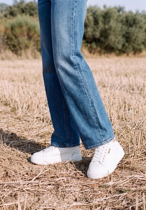 Gros plan sur une personne portant un jean bleu et des baskets blanches, debout dans un champ d’herbe sèche.