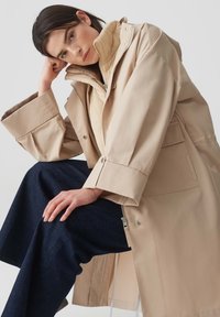 Cappotto impermeabile beige con colletto alto, caratterizzato da una chiusura con cerniera, maniche oversize e bottoni a pressione. Abbinato a pantaloni in denim scuri.