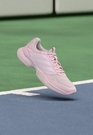 Chaussure de sport rose clair avec des rayures blanches flottant au-dessus d'une surface de court de tennis bleu et vert.