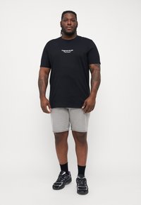 Sort bomulds t-shirt med et lille hvidt tekstlogo, parret med grå denimshorts og sorte sneakers med lilla accenter.