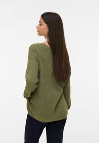 Vrouw met lang donker haar, gekleed in een losse groene trui en donkere broeken, staande met haar handen in de zakken, terwijl ze naar de zijkant kijkt tegen een witte achtergrond.