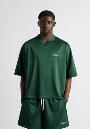 Homme debout devant un fond uni portant un maillot de sport en mesh vert foncé et un short assorti avec le logo "Buuoco" sur les deux.