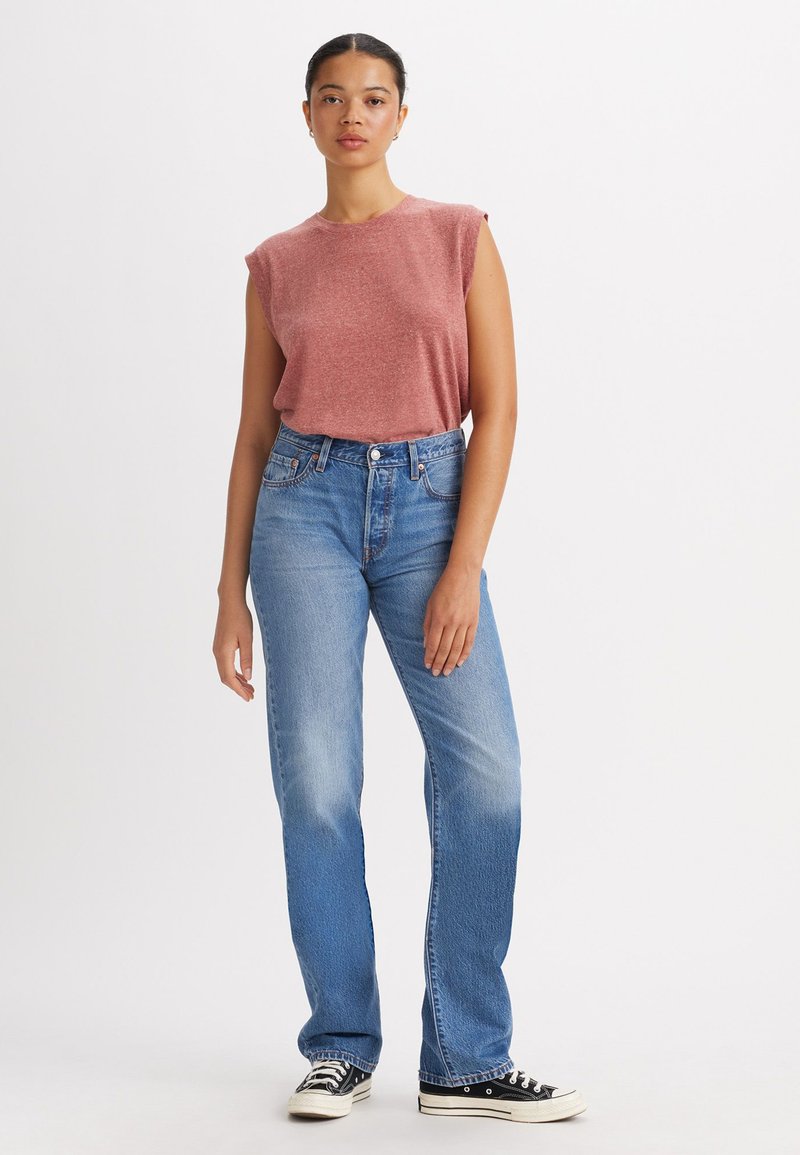 Rood heidelaar zonder mouwen gecombineerd met lichtblauwe rechtzittende jeans. Zwart-witte canvas sneakers maken de outfit af.