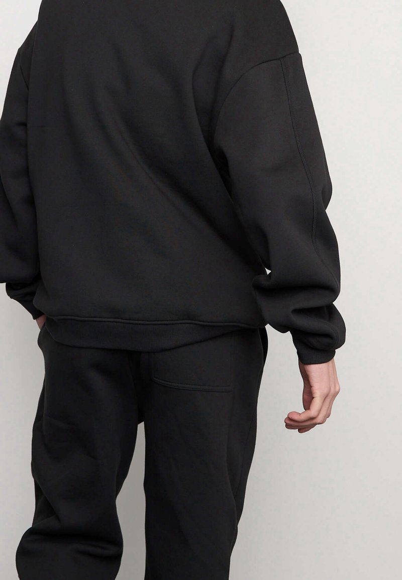 Maglione nero con pantaloni coordinati, caratterizzati da maniche lunghe, polsini a coste e una vestibilità rilassata con una tasca sul retro dei pantaloni. Texture liscia.