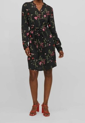 Czarna sukienka w kwiaty z długimi rękawami, dopasowaną talią i kołnierzykiem na guziki. Na materiale widoczne są różowe i zielone kwiaty. Połączona z czerwonymi sandałami na obcasie.