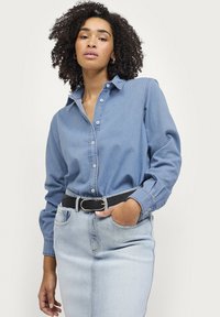 Lyseblå denimskjorte med lange ærmer, knaplukning foran og klassisk krave. Bæret med et sort bælte og lyse vaskede jeans.