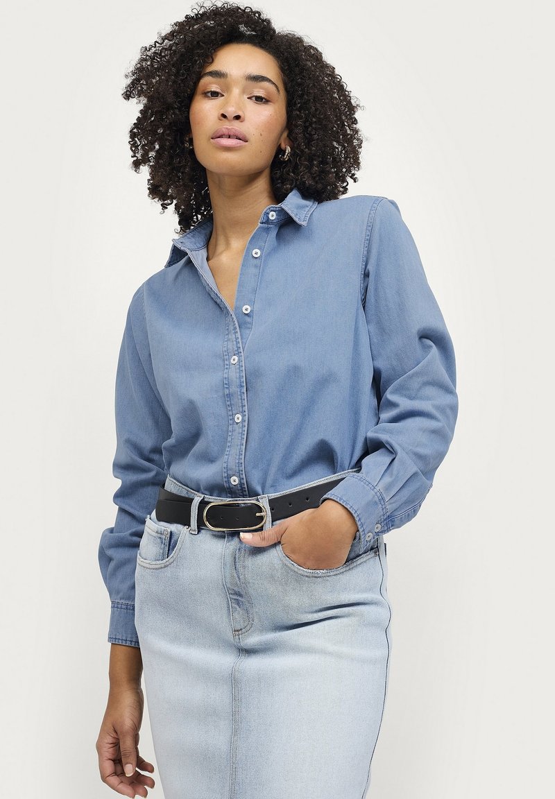 Lyseblå denimskjorte med lange ærmer, knaplukning foran og klassisk krave. Bæret med et sort bælte og lyse vaskede jeans.
