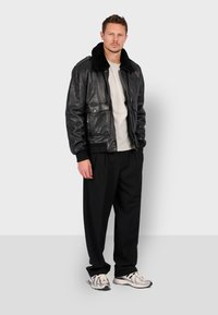 Černá kožená bomberka s ovčími límcem, dvěma předními kapsami, vroubkovanými manžetami, v kombinaci se šedým tričkem a volnými černými kalhotami.