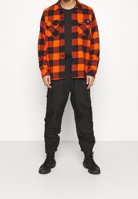 Camicia di flanella a quadri rossi e neri con due tasche sul petto, abbinata a pantaloni cargo neri e stivaletti neri con lacci. Outfit semplice e casual.