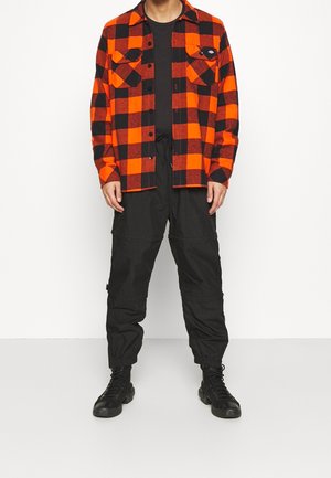 Camicia di flanella a quadri rossi e neri con due tasche sul petto, abbinata a pantaloni cargo neri e stivaletti neri con lacci. Outfit semplice e casual.
