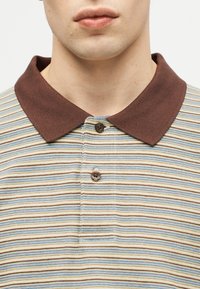 Κοντινό πλάνο ενός ατόμου που φοράει πουκάμισο πόλο με μπεζ ρίγες σε μπλέ και καφέ αποχρώσεις και καφέ μονόχρωμο γιακά, κουμπωμένο στην κορυφή.