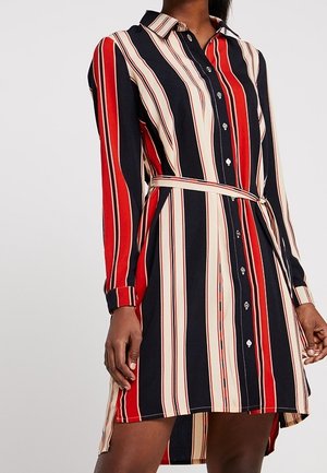 Femme portant une robe-chemise ceinturée à manches longues avec des rayures verticales rouges, noires et beiges, et une fermeture à boutons sur le devant.