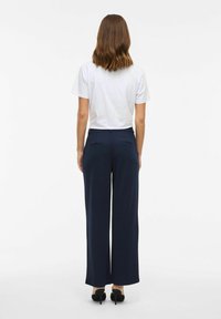 Vrouw met schouderlang bruin haar staat met haar rug naar ons toe, gekleed in een witte shirt met korte mouwen, marineblauwe wijde broek en zwarte hakken.
