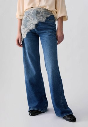 Person trägt weite blaue Jeans mit weißem Spitzen-Detail an einer Hüfte, beigefarbenes Langarmshirt und schwarze Schuhe, steht vor einem einfarbigen Hintergrund.