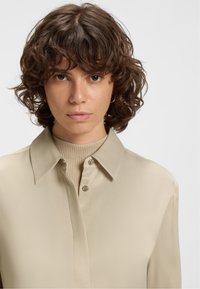 Woman with curly brown hair wearing gold hoop earrings, a beige collared shirt over a ribbed knit top, facing forward with a neutral expression.