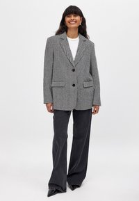 Harmaa houndstooth-takki, jossa on kaksi etutaskua, yksittäinen napitus, viistot läpät ja rento istuvuus. Yhdistetty tummiin leveisiin farkkuihin.