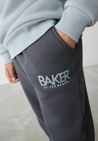 Grå sweatsbukser med et logo, der står "BAKER by Ted Baker" i lys grå. Bukserne har et blødt, tekstureret stof og sidelommer.