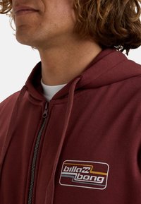 Sudadera con cremallera en color marrón con capucha ajustable mediante cordones, que presenta un parche de logo blanco y multicolor en el lado izquierdo del pecho. Tejido suave y texturizado.