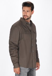 Donkerbruine blouse met lange mouwen en knoopsluiting, twee borstzakken, klassieke kraag en een soepele stofstructuur. G gedragen met grijze jeans.