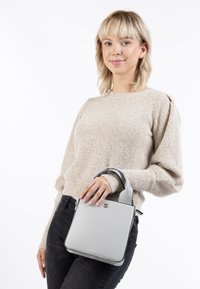 Borsa quadrata grigia realizzata in pelle liscia con un design strutturato, manici corti e un'emblema in metallo sulla parte anteriore.