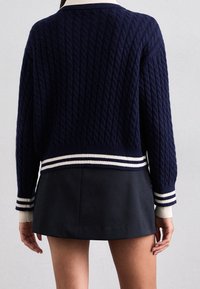 Marinblå kabelstickad tröja med en vit randig ribbad nederkant och ärmslut. Den har en rund halsringning och en avslappnad passform, ihop med en mörk kjol.
