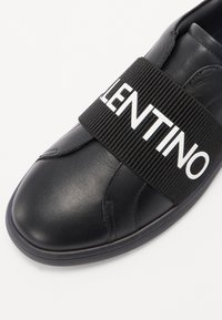 Sneaker in pelle nera con una spessa fascia elastica nera che presenta "VALENTINO" in lettere bianche a metà piede. Texture liscia, punta rotonda.