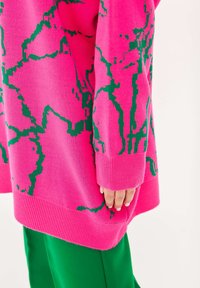 Rosa stickad tröja med grönt abstrakt mönster, ribbade ärmar och lös passform, kombinerad med gröna byxor, som visar en hand på sidan.