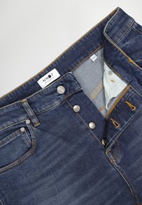 Jeans de mezclilla azul oscuro con cierre de botón, costuras naranjas, bolsillos frontales y una etiqueta blanca que dice "NN07 No Nationality" en el interior de la cintura.