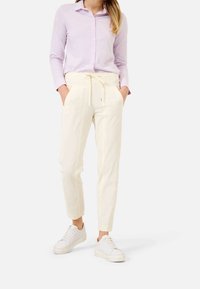 Kobieta ubrana w jasnofioletową koszulę na guziki, kremowe spodnie na sznurku i białe trampki, stoi z rękami w kieszeniach na białym tle.