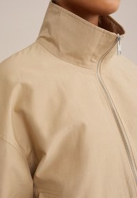 béžová lehká bunda s vysokým límcem, stříbrným zipem a hladkým, mírně texturovaným materiálem. Má minimalistický design.