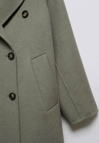 Manteau en laine vert olive avec devant croisé, grands boutons noirs et poche latérale. Texture douce et coupe décontractée.