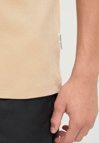 Beige cotton t-shirt with short sleeves, side label reading "FRIDAY CASUAL," worn with black pants. Smooth texture and fitted design.
