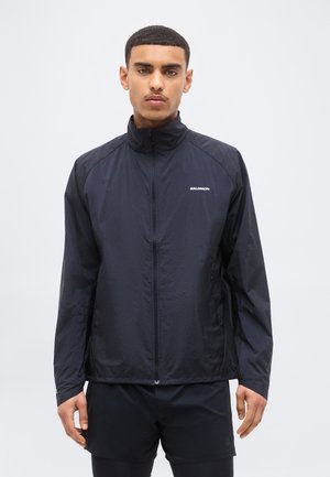 Άνδρας που φοράει μαύρο ελαφρύ μπουφάν Salomon με φερμουάρ και μαύρο σορτς, στέκεται μπροστά σε απλό φωτεινό φόντο.