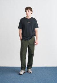 T-shirt preta de manga curta com um pequeno logótipo, combinada com calças cargo verde-oliva e ténis cinza claro, em pé num tapete azul.